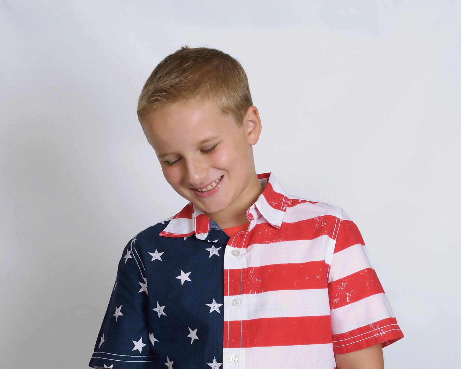 A smiling boy in a stars-and-stripes shirt, looking down with a grin